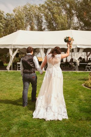 A celebratory outdoor wedding scene with a couple raising their hands beneath a white tent on a grassy lawn.