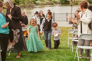 Children in formal attire walk among wedding guests seated on white folding chairs at an outdoor ceremony.