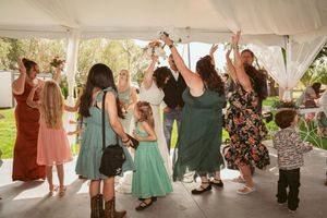 A group of wedding guests dancing and celebrating under a white outdoor tent at a daytime reception.