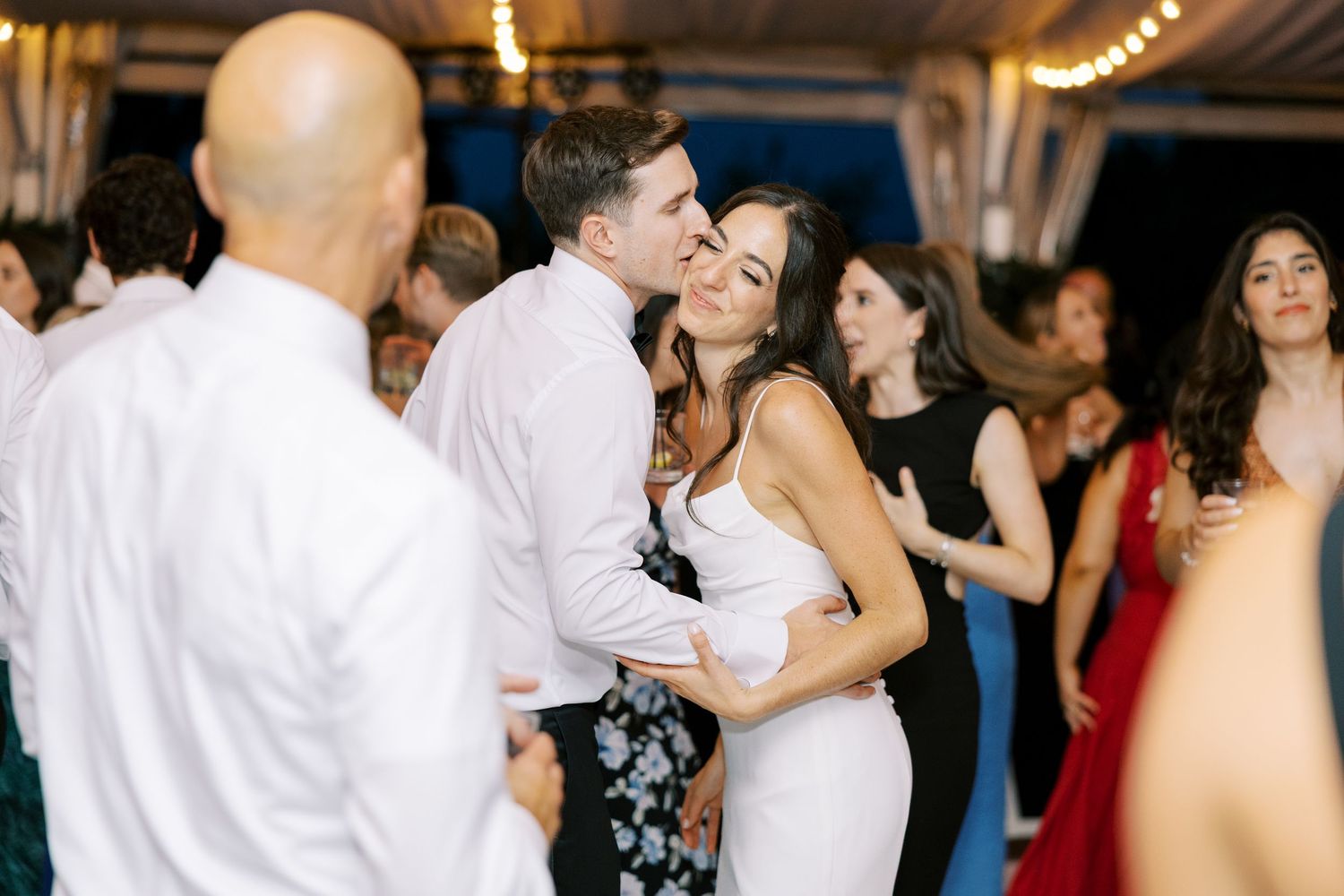 Couples dance together at an indoor wedding reception with string lights overhead.