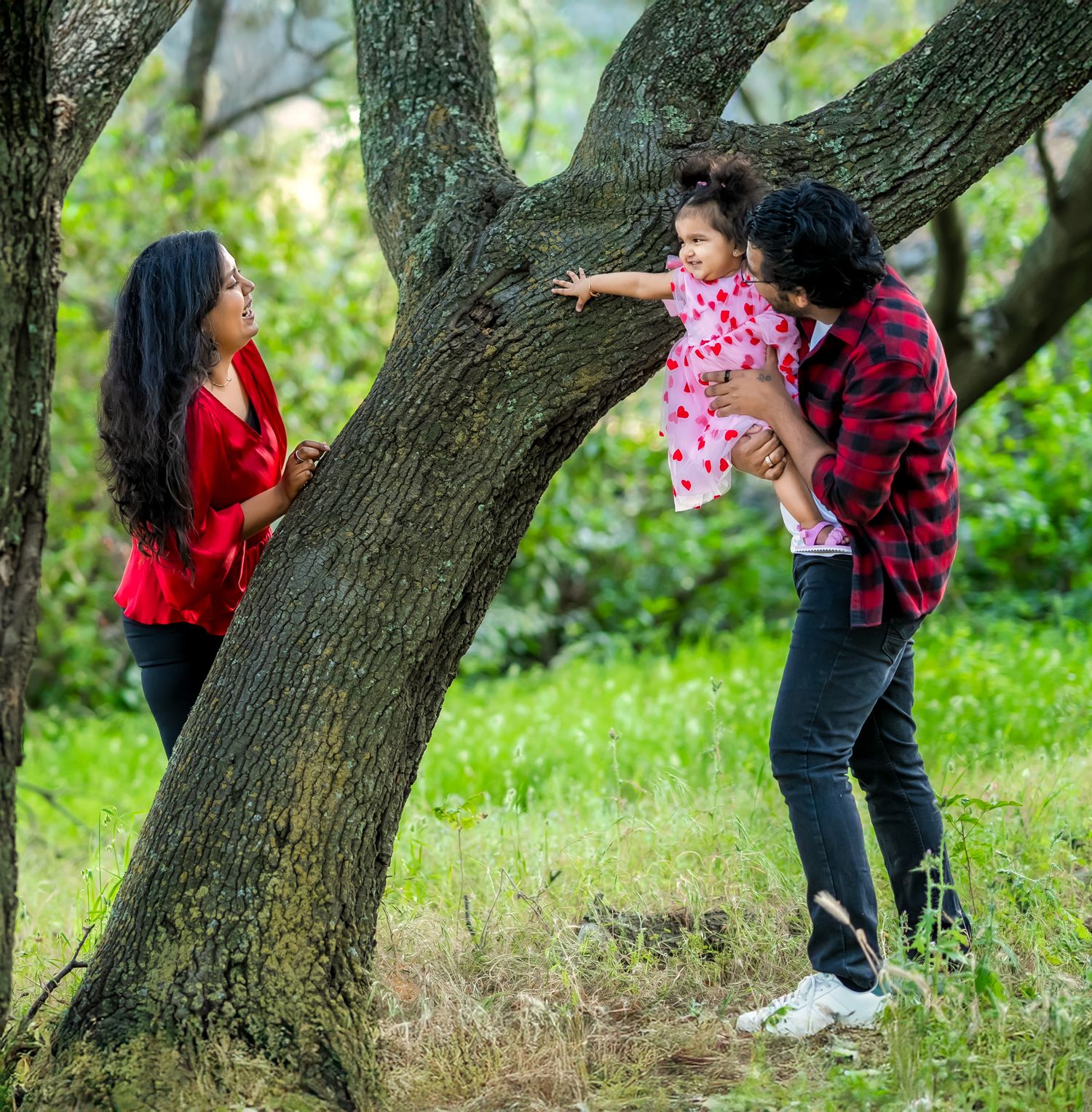 Couple in matching red plaid shirts enjoy a playful moment under a large tree in a sunny green park.