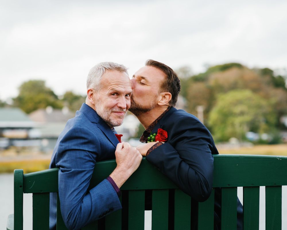 Two people in formal attire sitting on a green bench outdoors share an affectionate moment together.