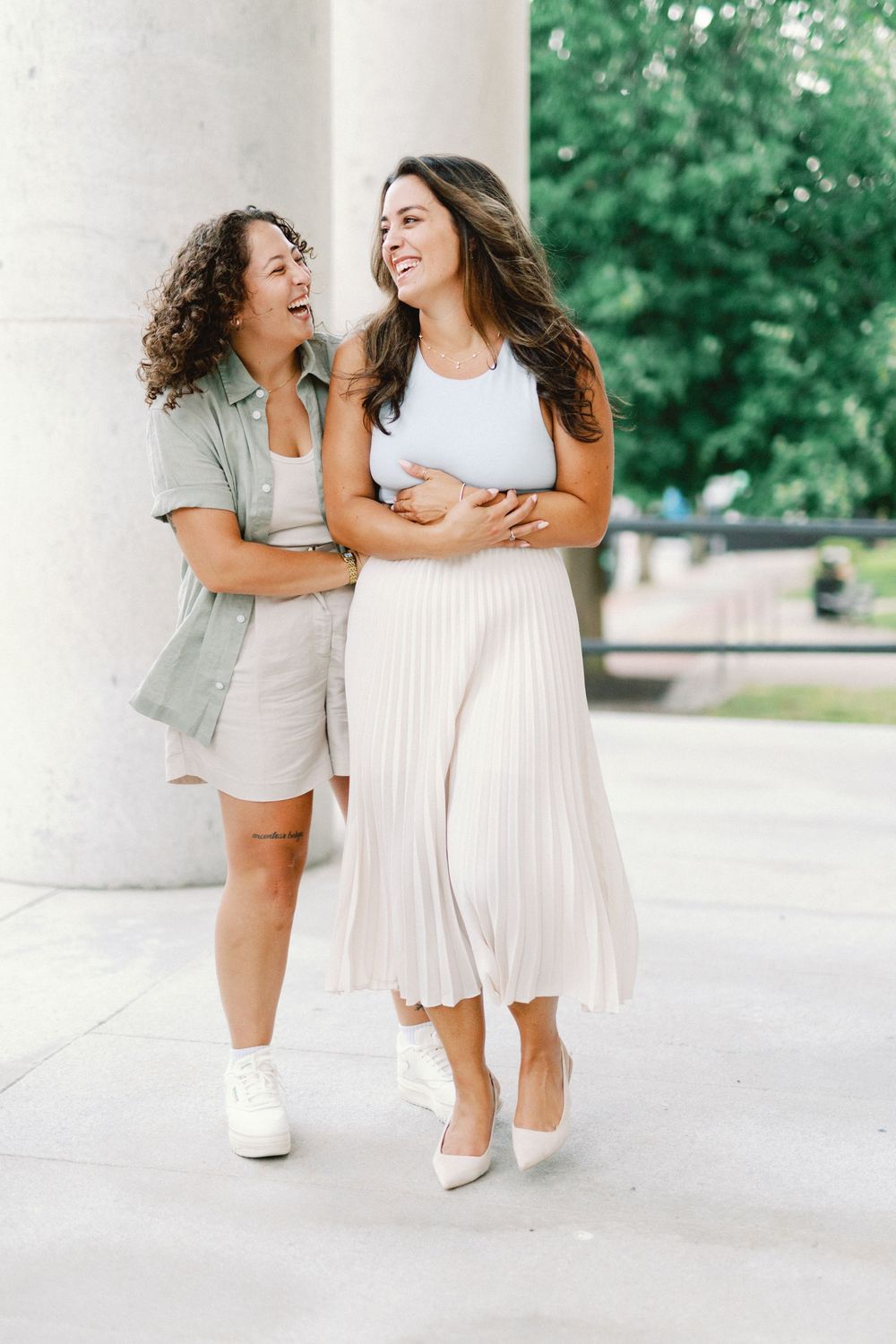 Two friends laugh together while walking outdoors, one wearing a pleated white skirt and the other in casual shorts.