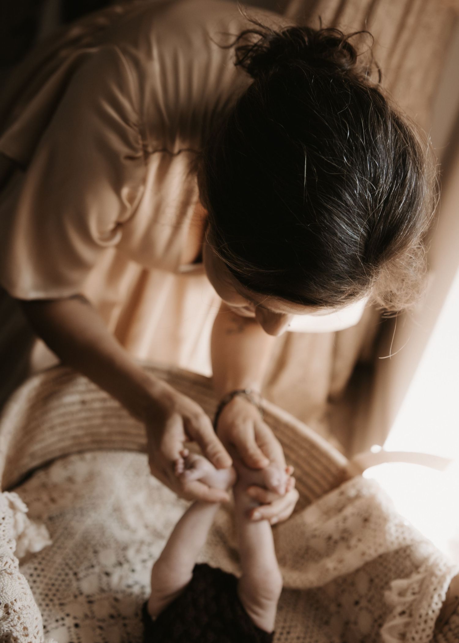 A mother tenderly holds her newborn baby's feet while leaning over a wicker bassinet in warm natural light.