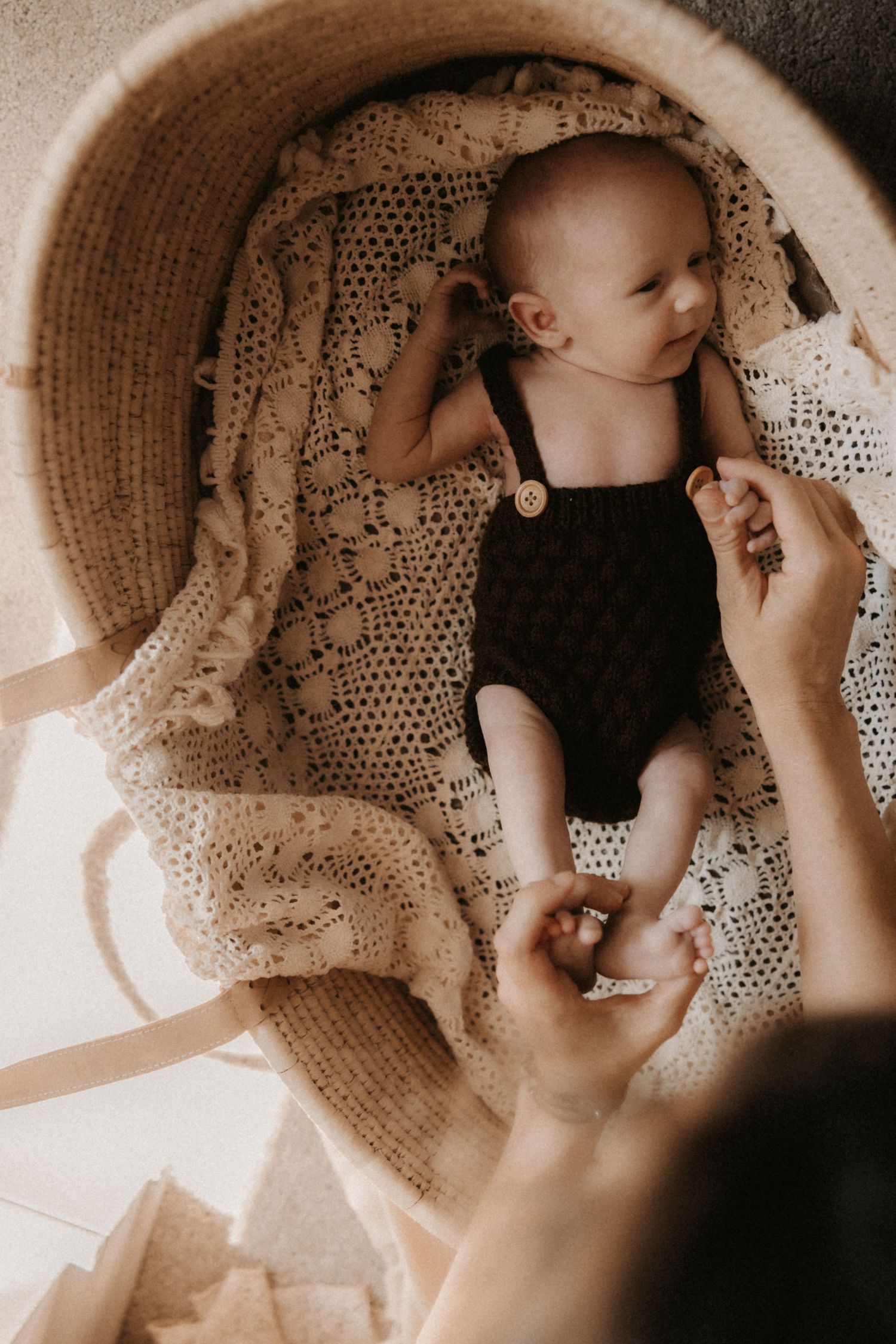Newborn baby in dark knit romper lying in wicker Moses basket with crochet blanket, parent holding tiny feet.