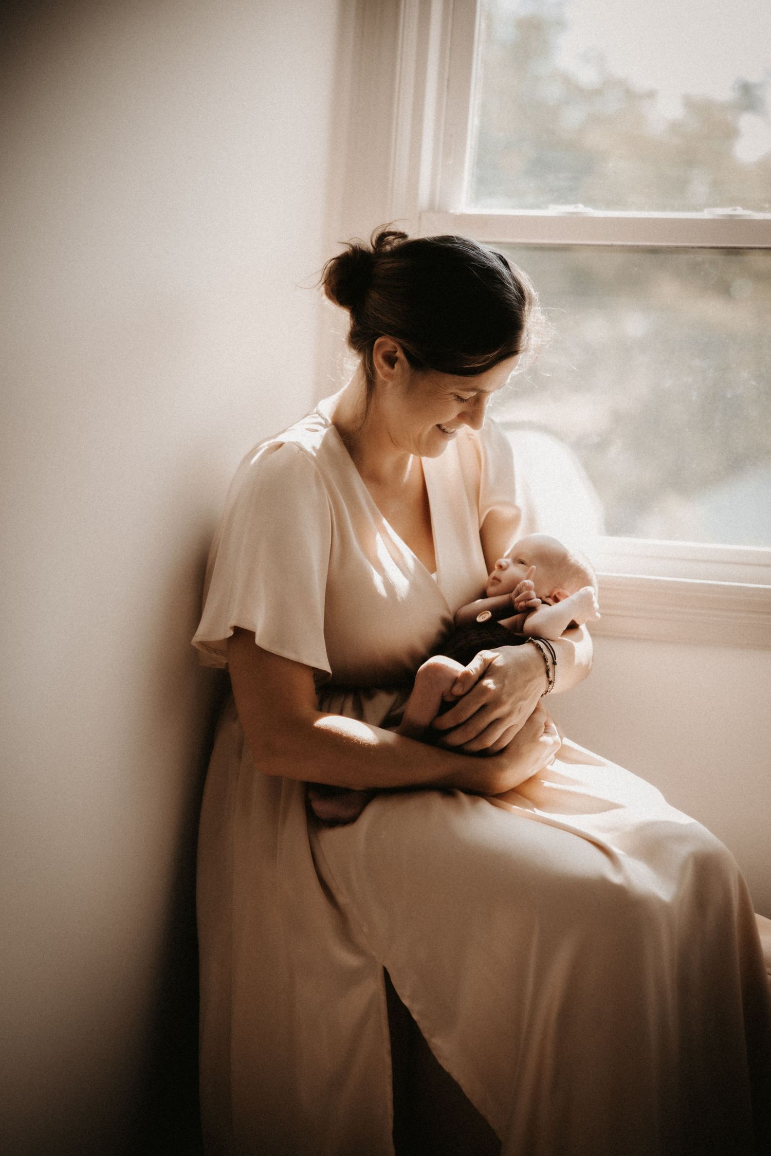 A mother in a white dress sits by a sunlit window, tenderly cradling and breastfeeding her newborn baby.