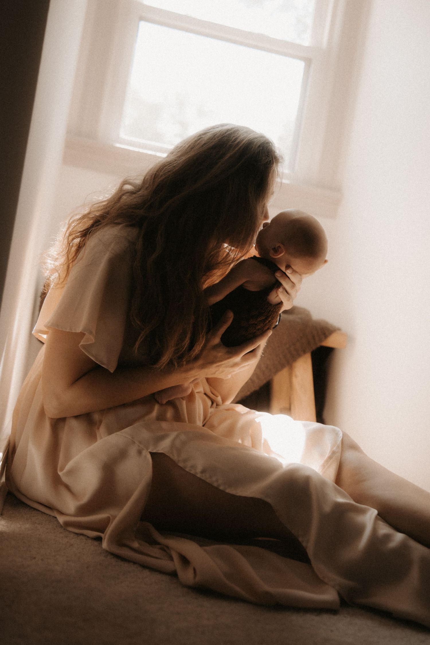 Mother in cream silk dress sitting by window, tenderly kissing her newborn baby in soft natural light.