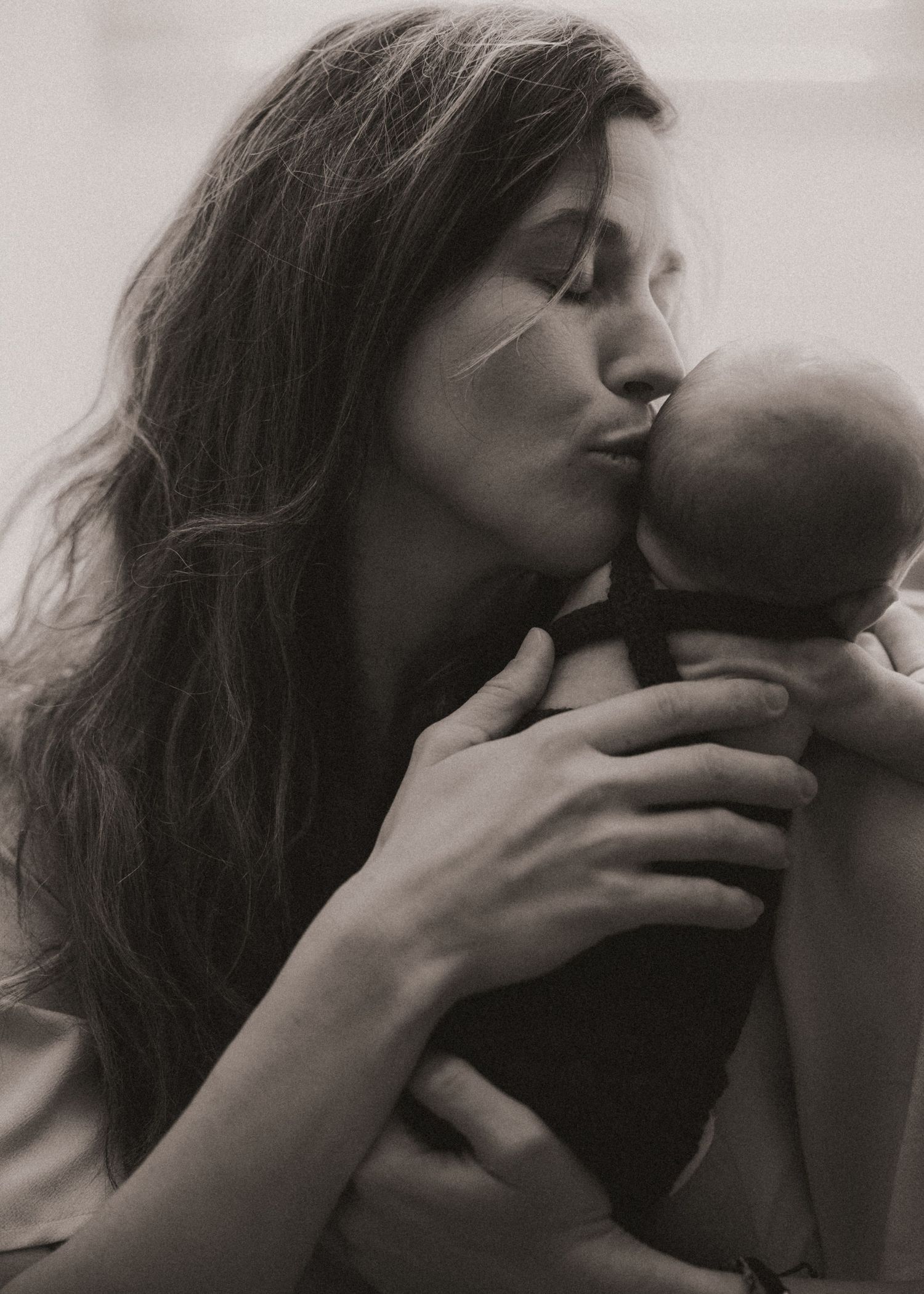 Black and white photo of a mother tenderly kissing her newborn baby while cradling the infant in her arms.