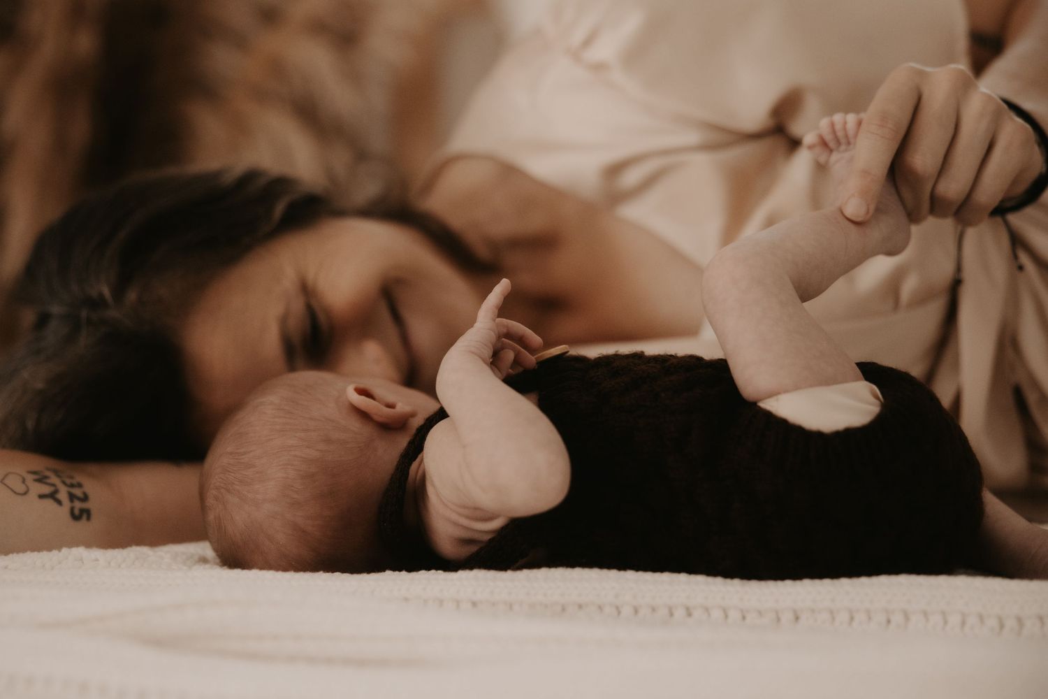 A mother lying on a white bed gazes lovingly at her newborn baby dressed in black.