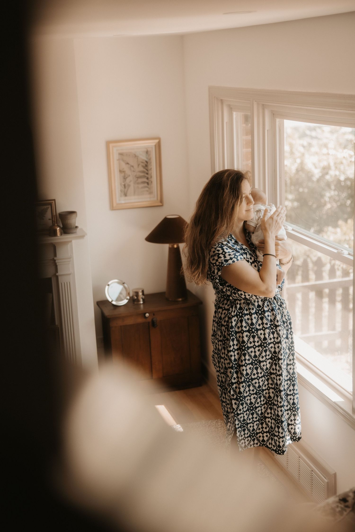 A woman in a floral dress holds her newborn near a sunlit window in a cozy home interior.