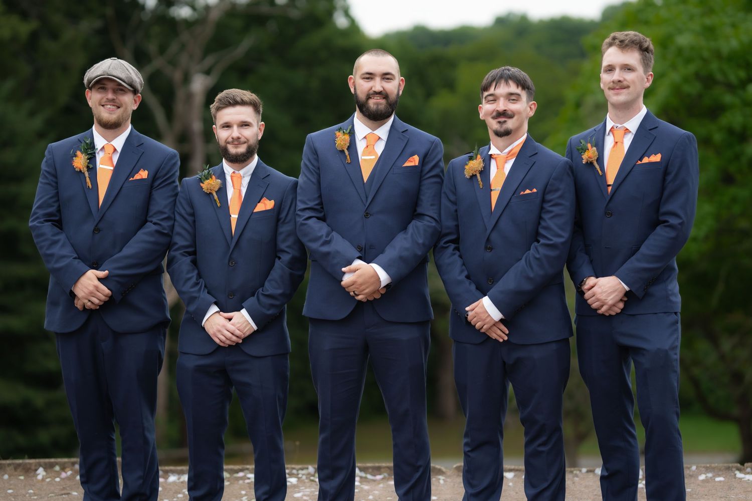 Five groomsmen in matching navy suits with orange accents pose formally outdoors.