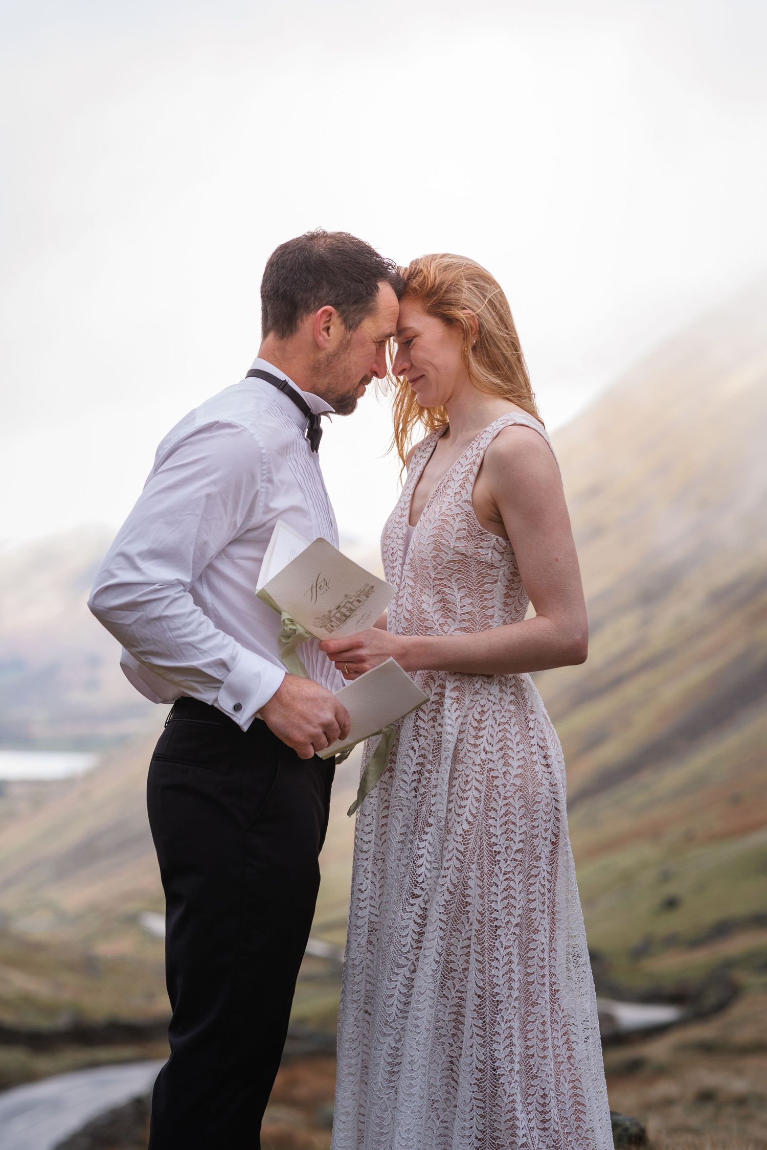 A couple shares an intimate moment in their wedding attire against a misty mountain backdrop in Scotland.