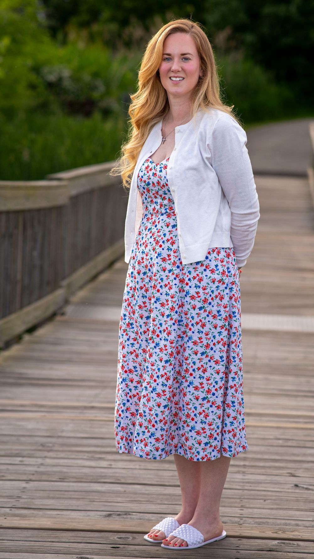 Woman in a white cardigan and floral skirt standing on a wooden boardwalk during a family portrait session.