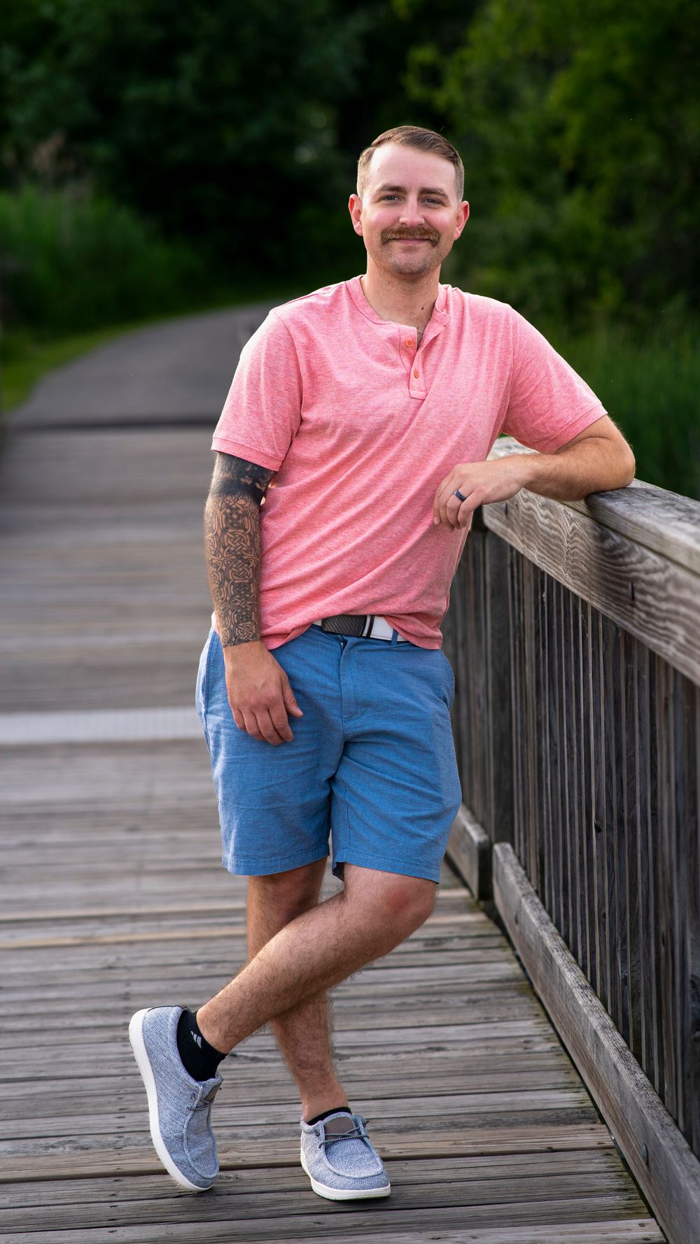 Man in a pink shirt and denim shorts standing on a wooden boardwalk during a portrait session.