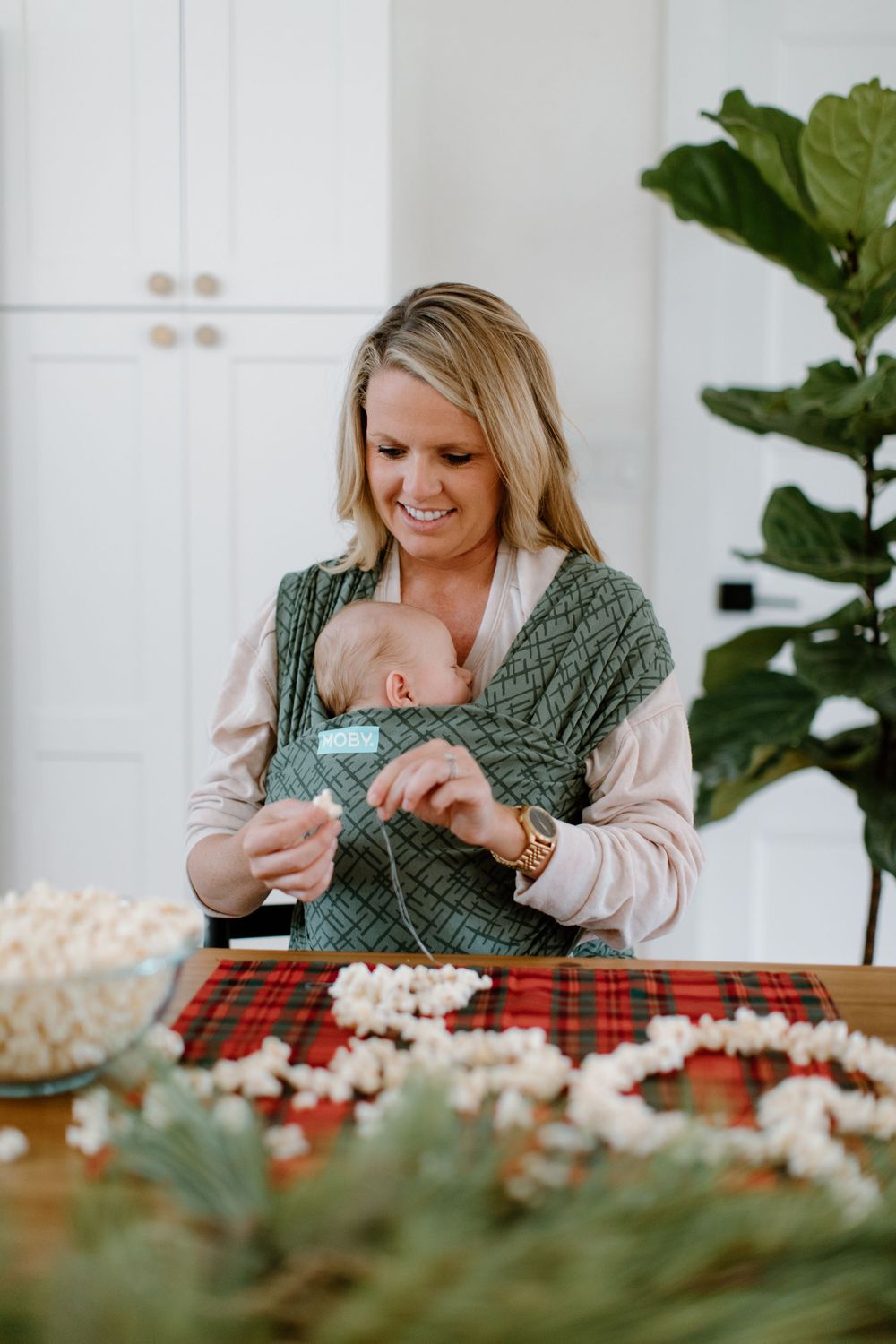 Baby wearing in a green carrier while decorating with popcorn garlands on a plaid holiday tablecloth.