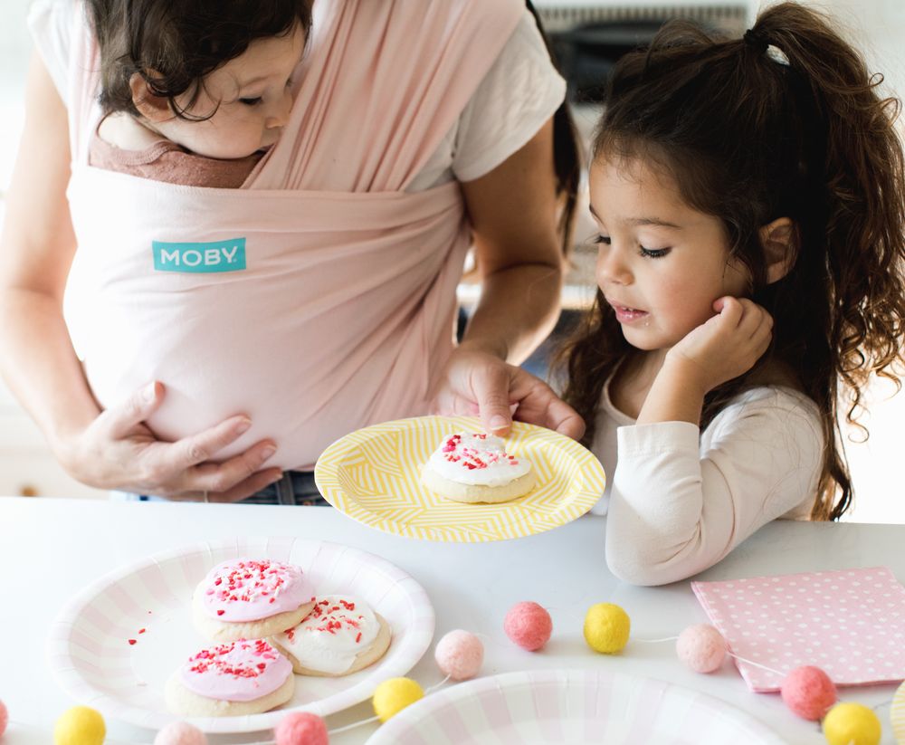 A baby in a pink carrier while a child decorates cookies at a table.
