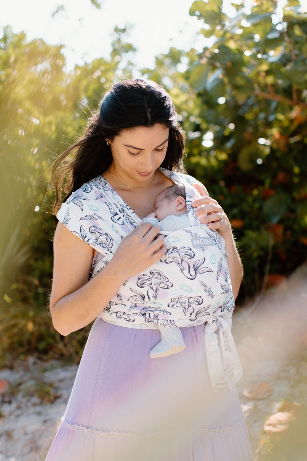 A baby being carried in a printed wrap carrier outdoors in golden sunlight.
