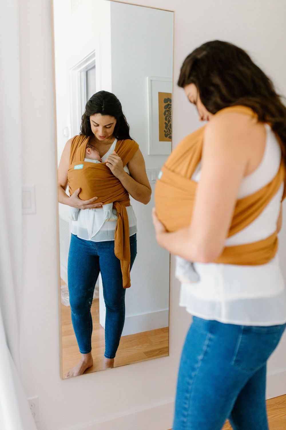 A parent adjusting a brown baby carrier wrap while looking in a full-length mirror.
