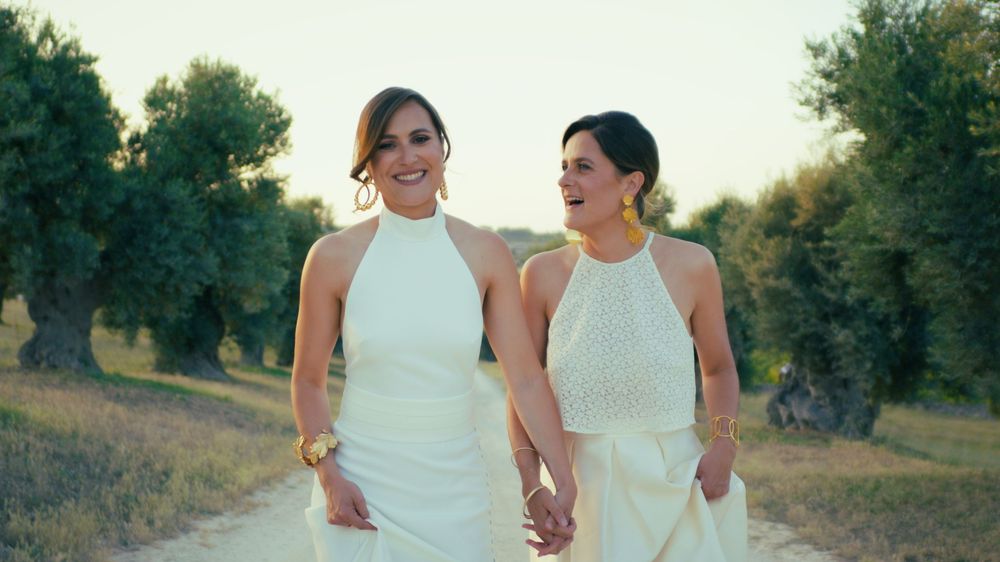 Two people in elegant white dresses walk hand in hand on a path with olive trees at golden hour.