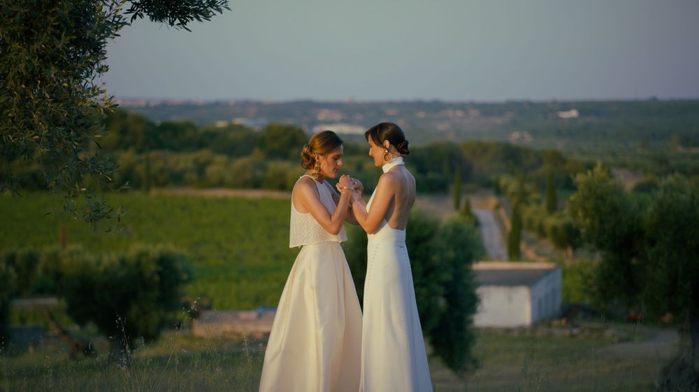 Wedding party members in cream-colored dresses gather on a scenic hilltop overlooking a countryside landscape at sunset.