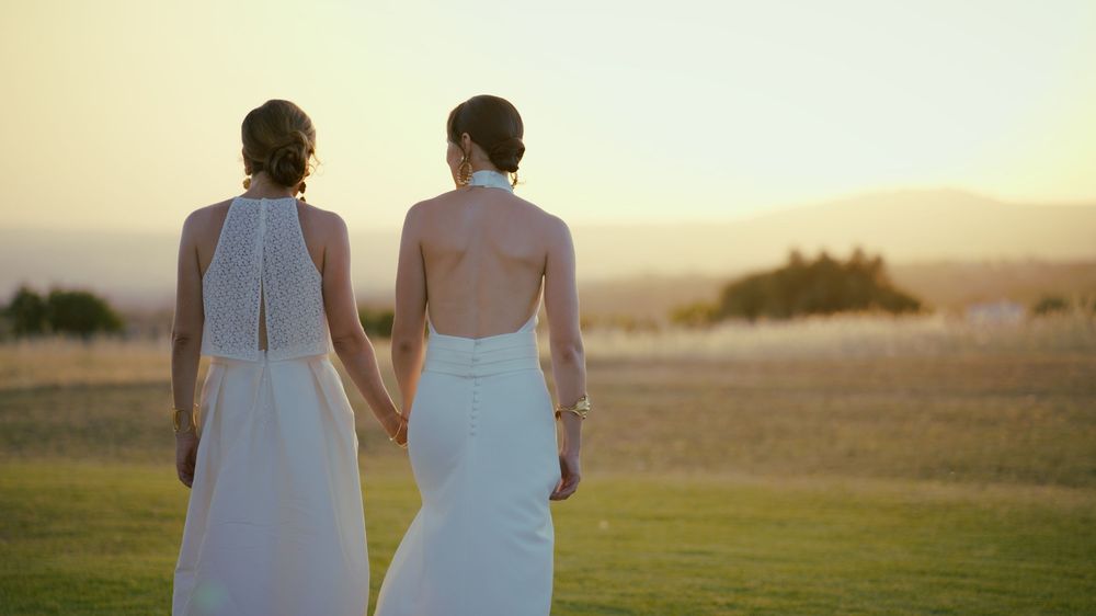 Couple holding hands walking together across golden fields at sunset with light rays glowing behind them.