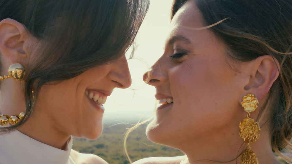 Two people share a romantic moment at sunset while wearing elegant gold jewelry and formal attire.