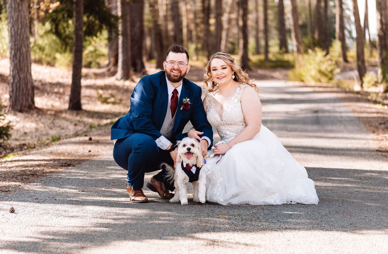 A couple in wedding attire poses together with their fluffy white dog on a pathway lined with tall pine trees.