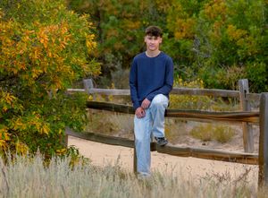 Someone in blue denim and a navy sweater sits casually on a wooden fence against vibrant autumn foliage.