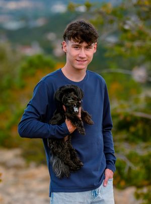 A person in a navy blue sweater holds a small black dog while standing outdoors in a natural setting.