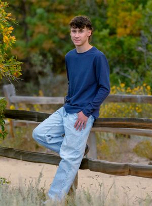 Person in a navy sweater and light jeans sits casually on a wooden railing during autumn.