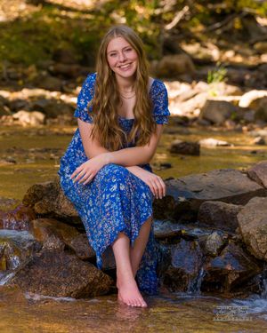 Someone in a blue floral dress sits barefoot on rocks by a stream in a sunlit forest setting.