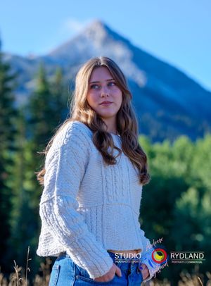 A person in a white cable knit sweater poses outdoors against a backdrop of majestic mountain peaks.
