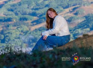 A person in casual attire sits peacefully on a hillside overlooking a scenic mountain valley at dusk.