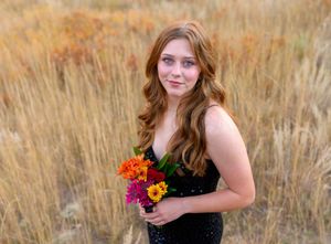 A woman in a black dress poses in a golden wheat field holding colorful flowers during an outdoor photo session.