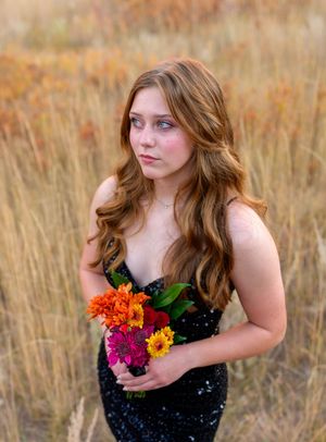 A woman in a black sequined dress poses with vibrant orange and pink flowers in a golden autumn field.
