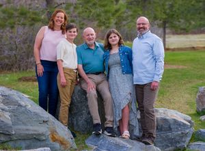A small group poses outdoors on a large boulder in a grassy area with trees in the background on a sunny day.