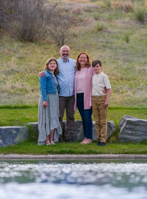 A group of four people stand together outdoors near a pond on grassy terrain with natural landscaping.