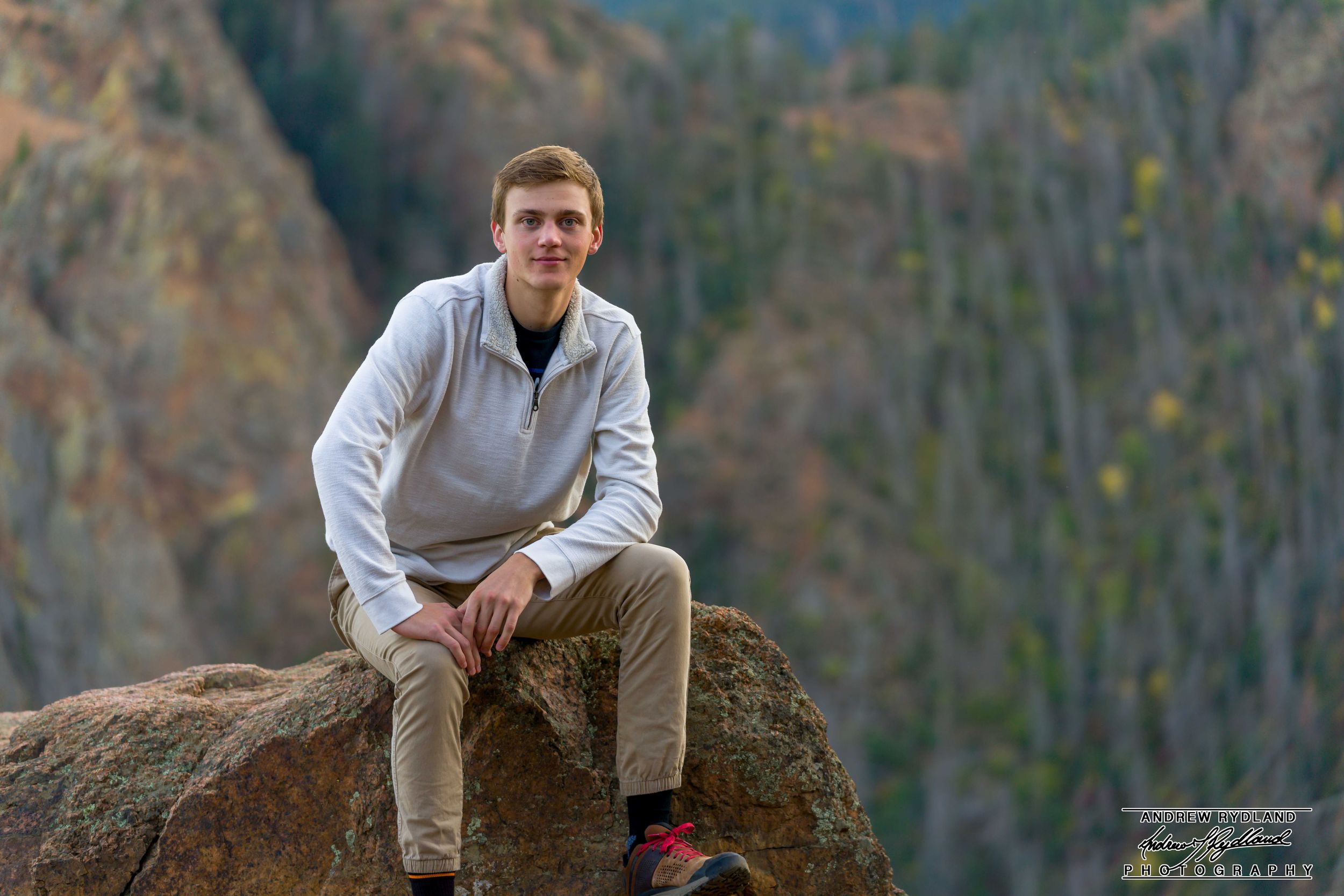 Someone in a light gray pullover and khaki pants sits on a large boulder with scenic mountain views in the background.