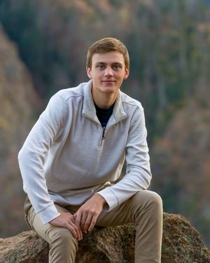 A person in a light gray quarter-zip sweater sits casually on a rocky overlook in a mountainous outdoor setting.