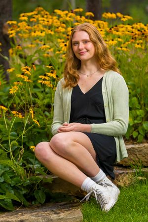 A person sits in a black dress and light cardigan among vibrant yellow wildflowers blooming in a garden setting.