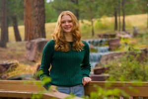 Young woman in green sweater and jeans poses outdoors in a scenic forest setting with evergreen trees and sunlight.