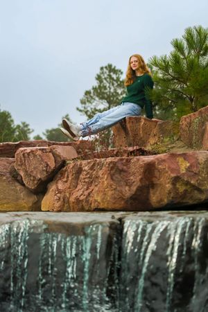 Someone in casual clothes sits on large red rocks near a waterfall while posing for a photo outdoors on a cloudy day.