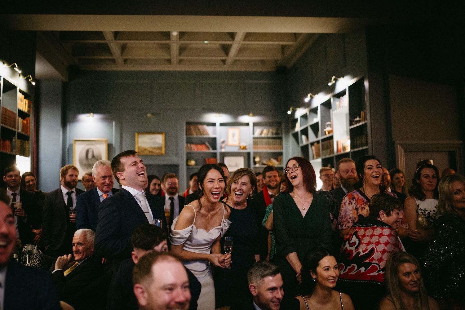 Guests laugh and smile while packed together in a dimly lit room with wooden shelves and artwork on the walls.