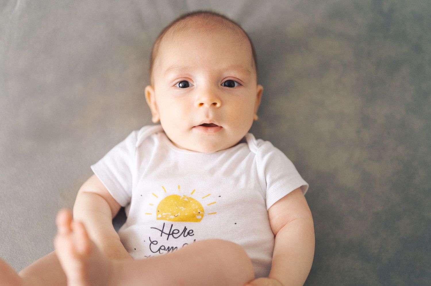 Baby wearing white onesie with yellow design lies on soft gray background.