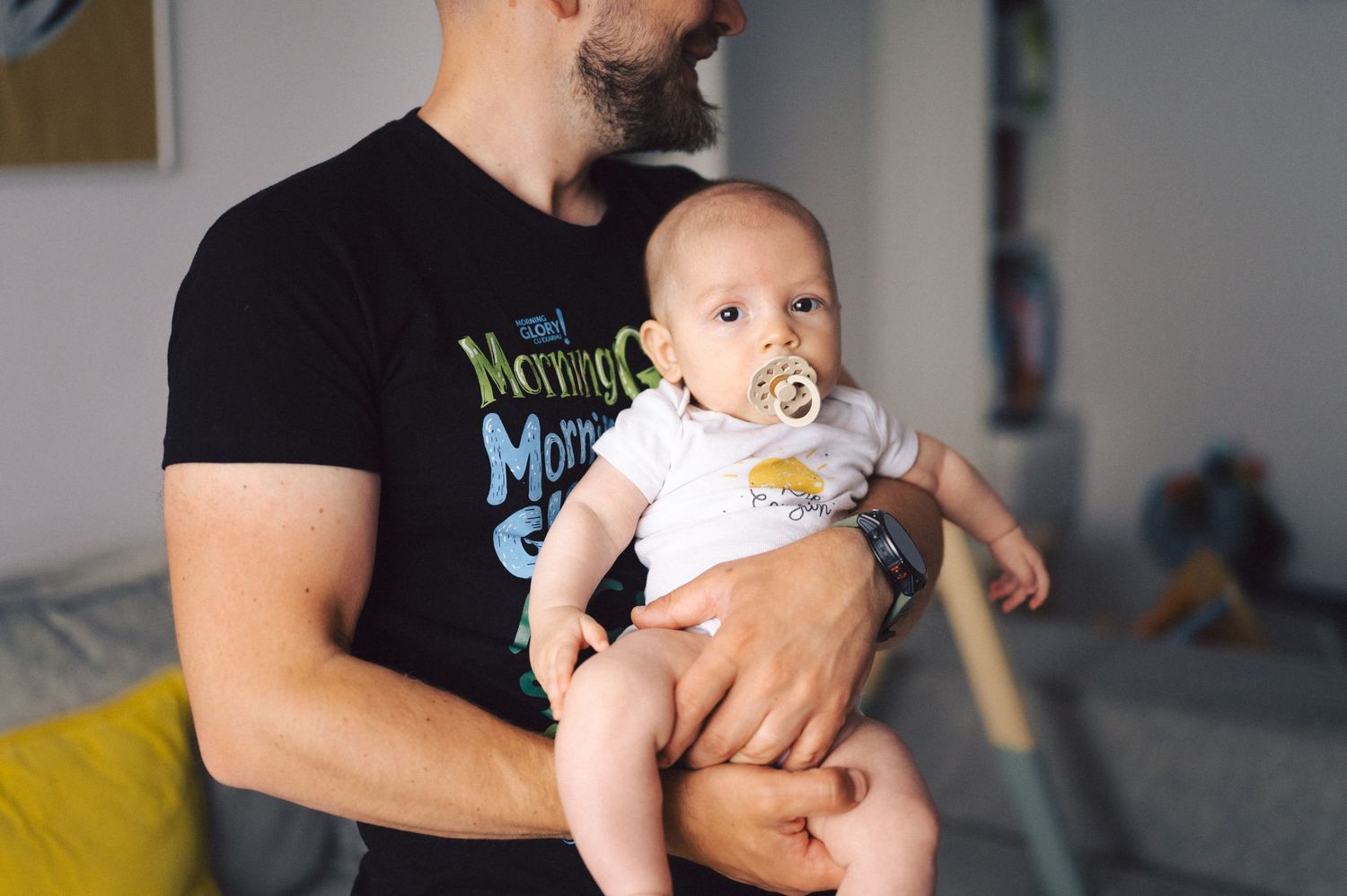 A baby wearing a white onesie and pacifier is held by someone in a black t-shirt.