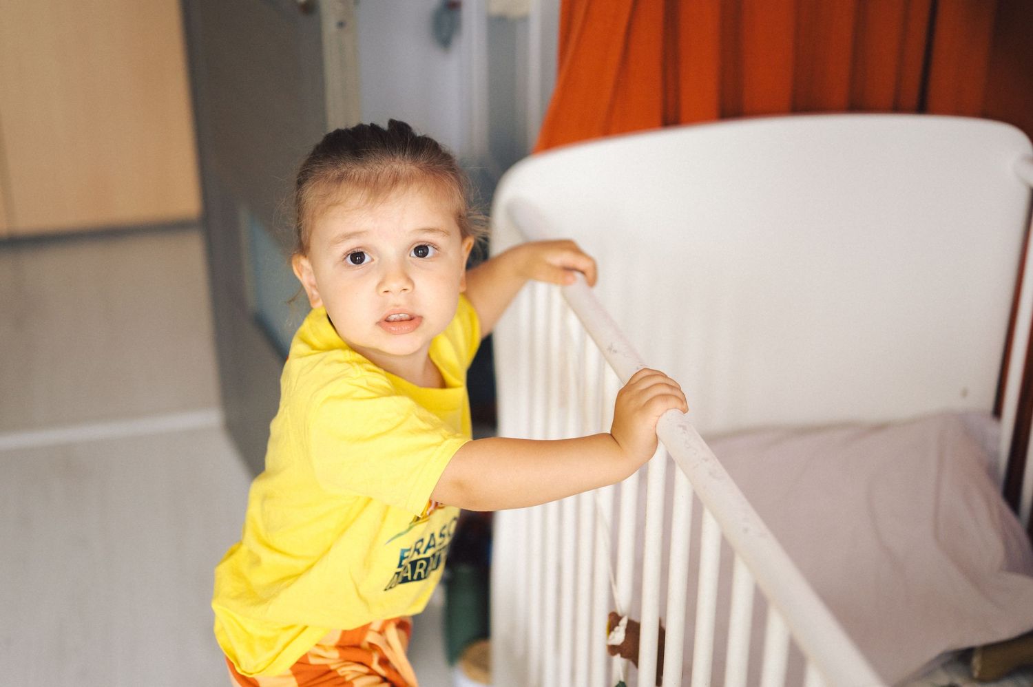 A small child wearing a bright yellow t-shirt stands near a white crib in a room.