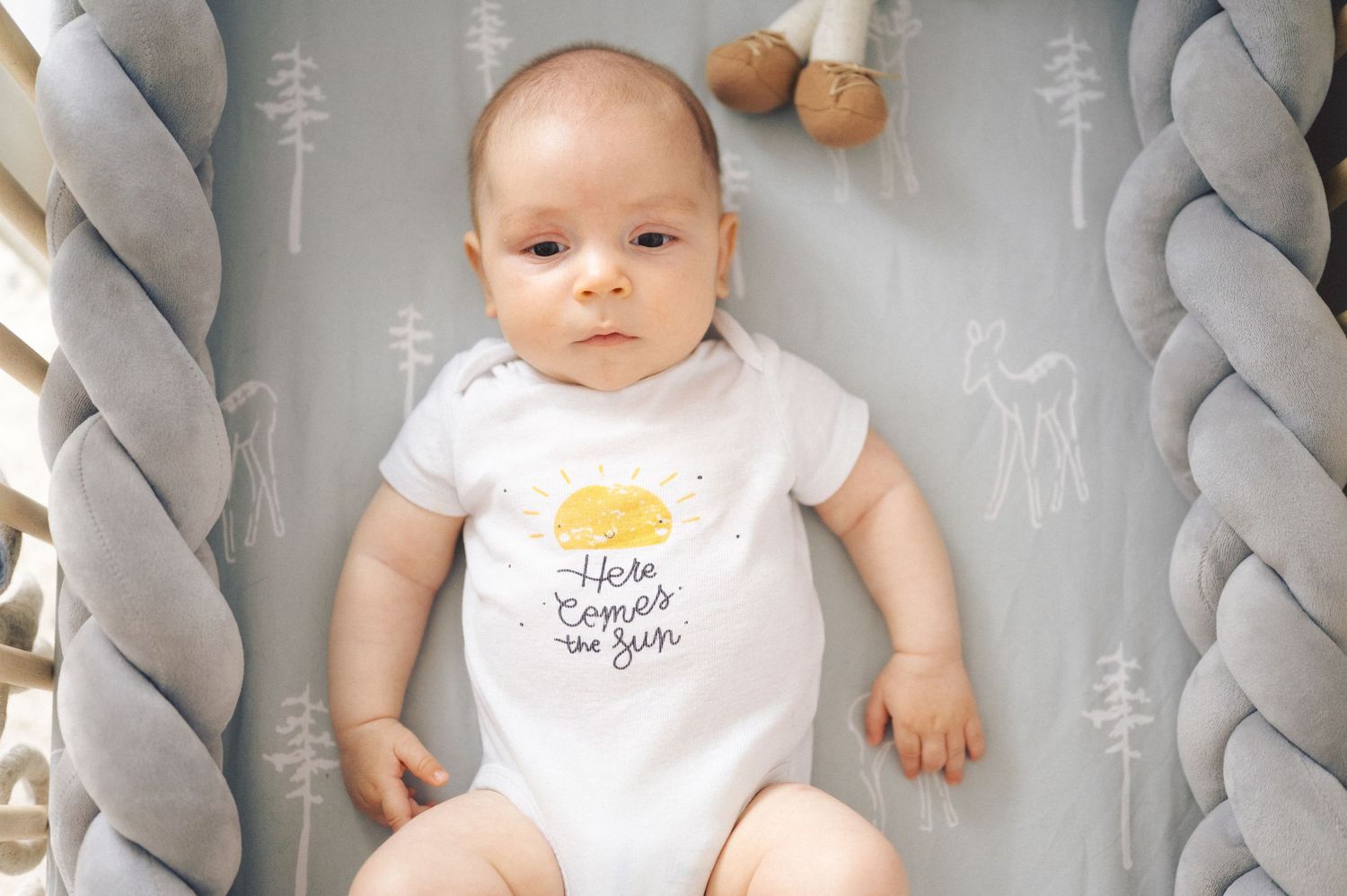 Baby wearing white onesie with sunny design lying on gray bedding with braided bolster cushions.