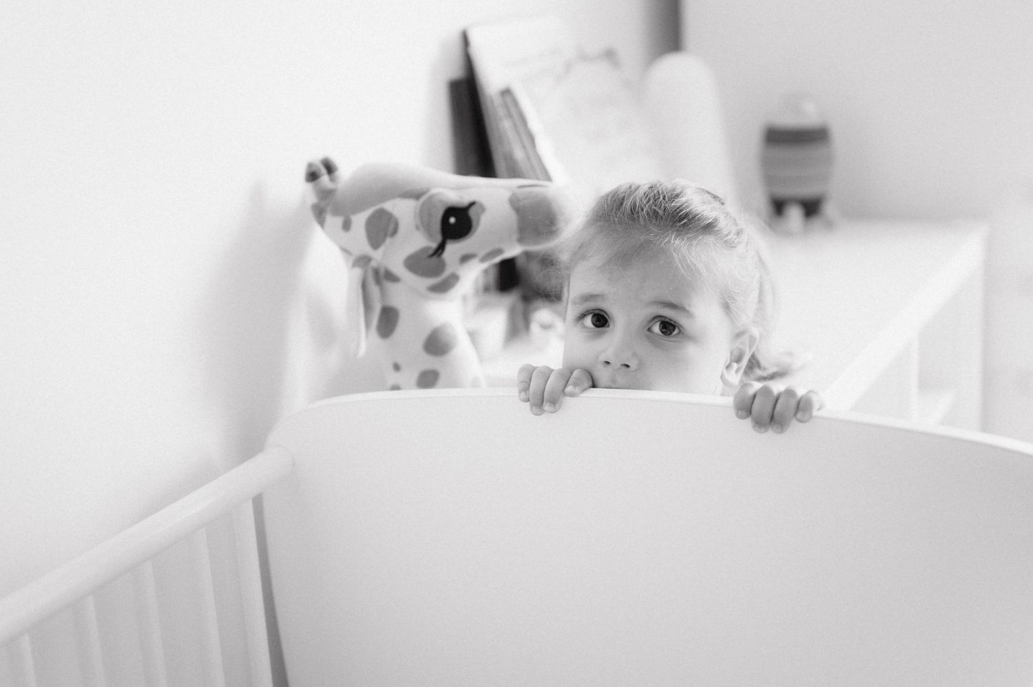 A child peeks over the edge of a white bed with wide eyes in this black and white photograph.