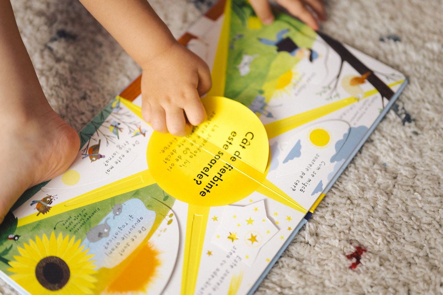 Small hand touches a yellow sun shape on an open children's book with colorful illustrations on a carpeted floor.