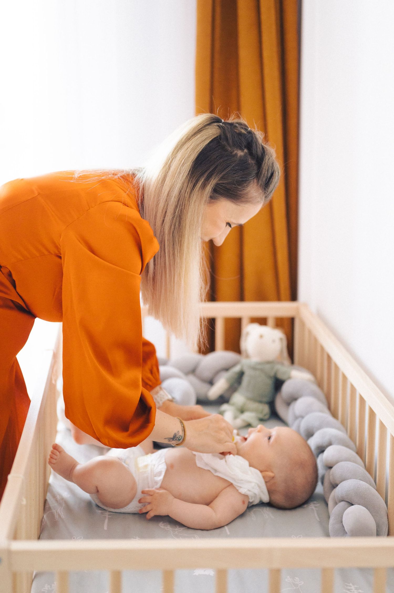 Parent in orange sweater tending to baby lying in wooden crib with plush gray bumper and stuffed toy.